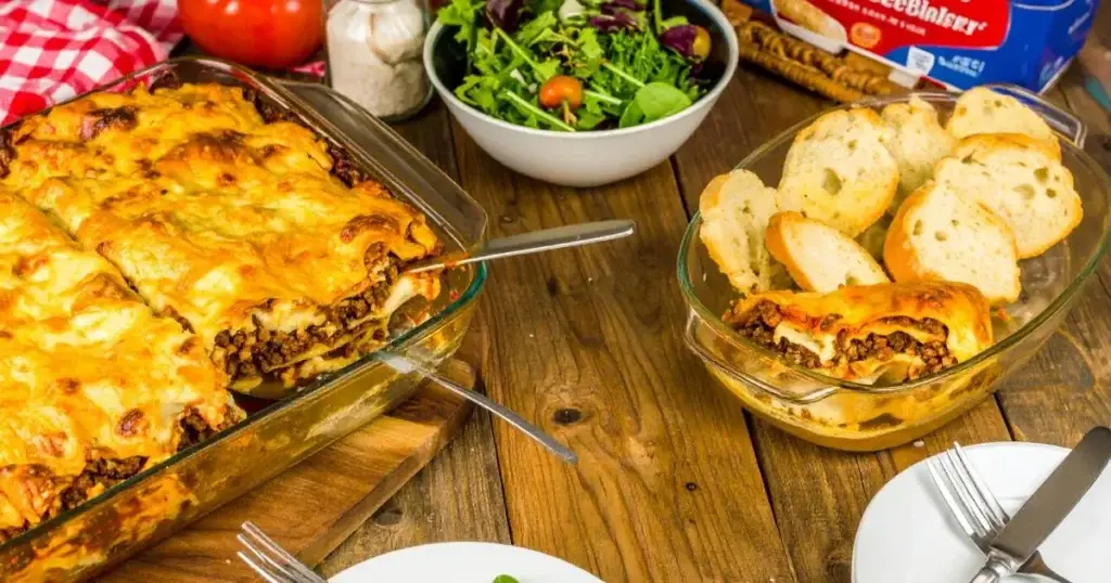 Freshly baked American Beauty lasagna recipe in a glass dish with golden melted cheese, visible layers of noodles, meat sauce, and ricotta, garnished with fresh basil, served on a rustic kitchen table with salad and garlic bread.