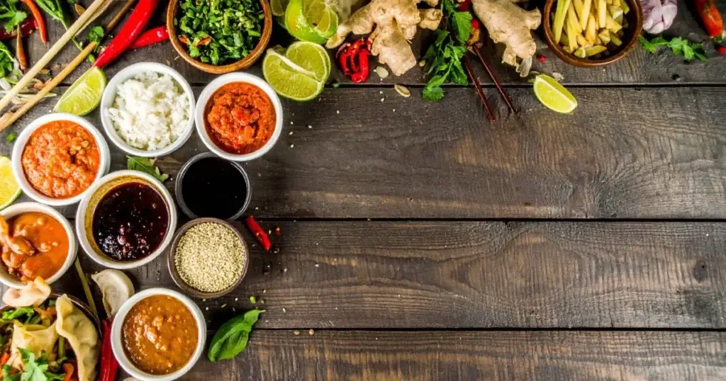 An overhead shot of assorted homemade Asian food sauces in small bowls, surrounded by fresh ingredients like ginger, garlic, chili peppers, and lime, with chopsticks and Asian dishes such as dumplings and spring rolls on a wooden table.