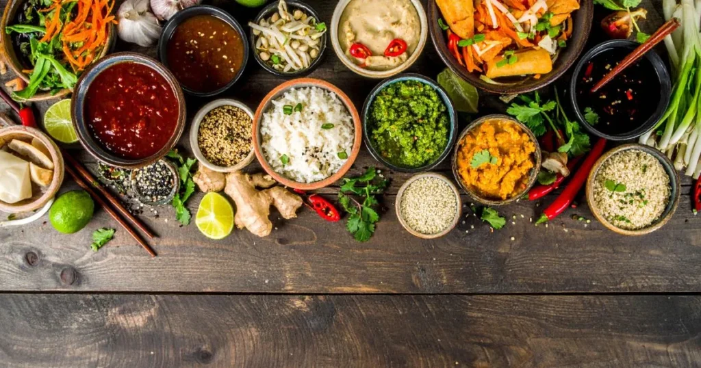 An overhead shot of assorted homemade Asian food sauces in small bowls, surrounded by fresh ingredients like ginger, garlic, chili peppers, and lime, with chopsticks and Asian dishes such as dumplings and spring rolls on a wooden table.