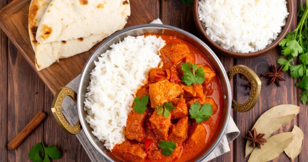 Lamb vindaloo recipe featuring tender lamb in spicy red curry, garnished with cilantro, served with rice and naan on a rustic wooden table.