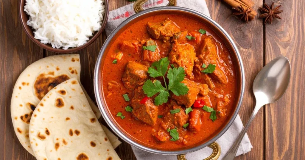 Lamb vindaloo recipe featuring tender lamb in spicy red curry, garnished with cilantro, served with rice and naan on a rustic wooden table.