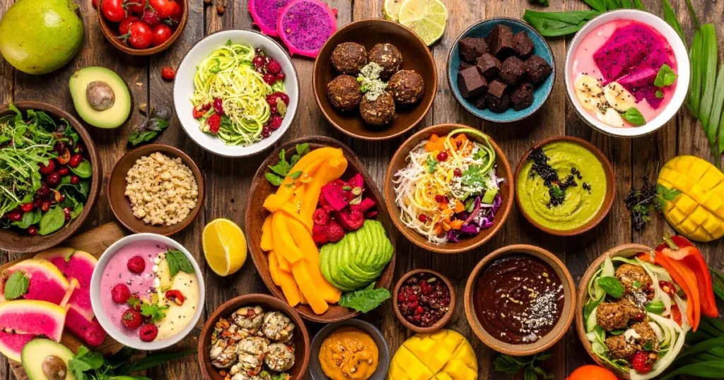 Colorful assortment of raw food recipes including zucchini noodles, avocado salad, smoothie bowls, and tropical fruits arranged on a wooden table — healthy, plant-based, and vibrant dishes.