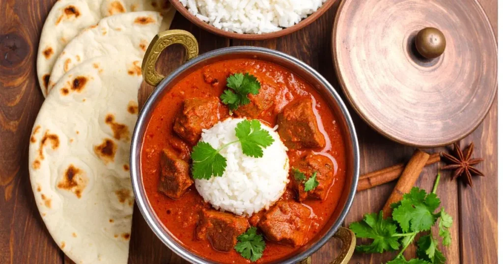 Lamb vindaloo recipe featuring tender lamb in spicy red curry, garnished with cilantro, served with rice and naan on a rustic wooden table.